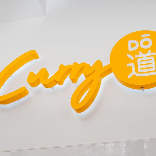 Bright yellow "Curry Dō" sign with Japanese characters, illuminated against a white wall.