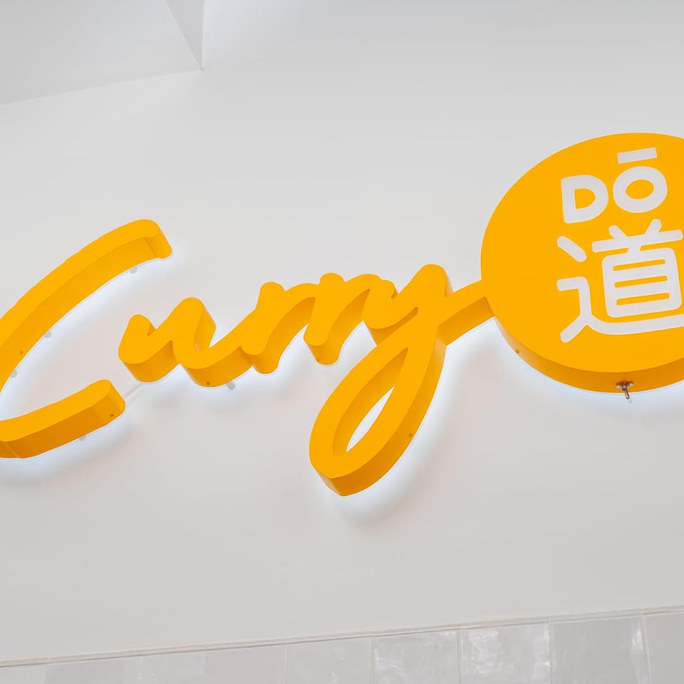Bright yellow "Curry Dō" sign with Japanese characters, illuminated against a white wall.