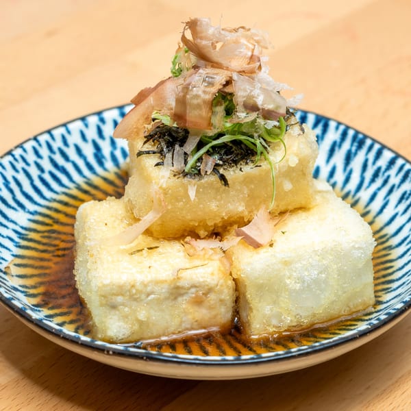 A close-up of agedashi tofu with bonito flakes, seaweed, and green onions in a patterned bowl.