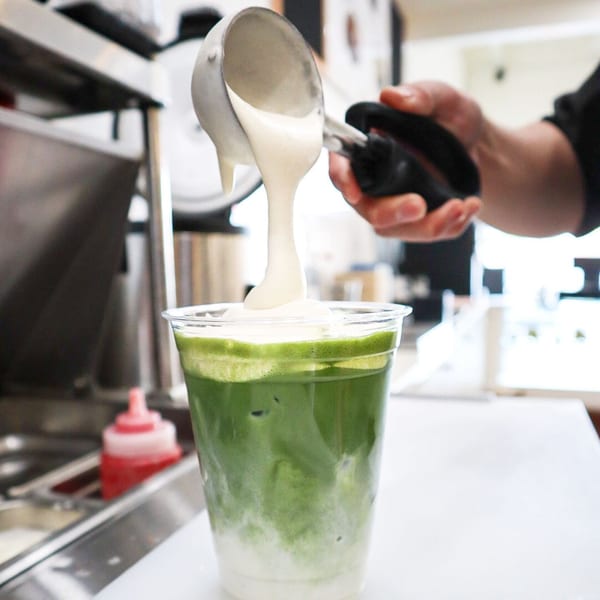 A hand pours creamy white foam from a ladle into a clear cup of green matcha latte.