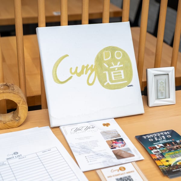 A white canvas with the gold "Curry Do" logo sits on a wooden table with flyers and a waitlist.