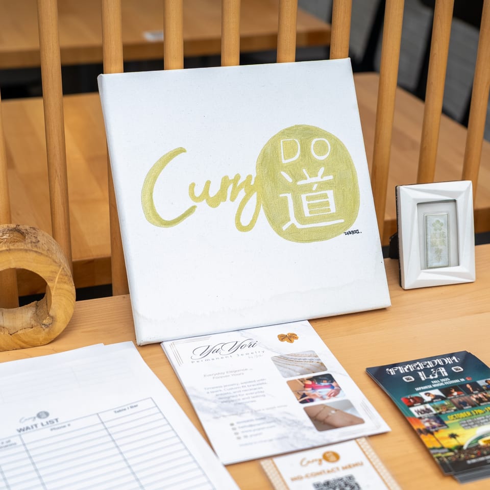 A white canvas with the gold "Curry Do" logo sits on a wooden table with flyers and a waitlist.