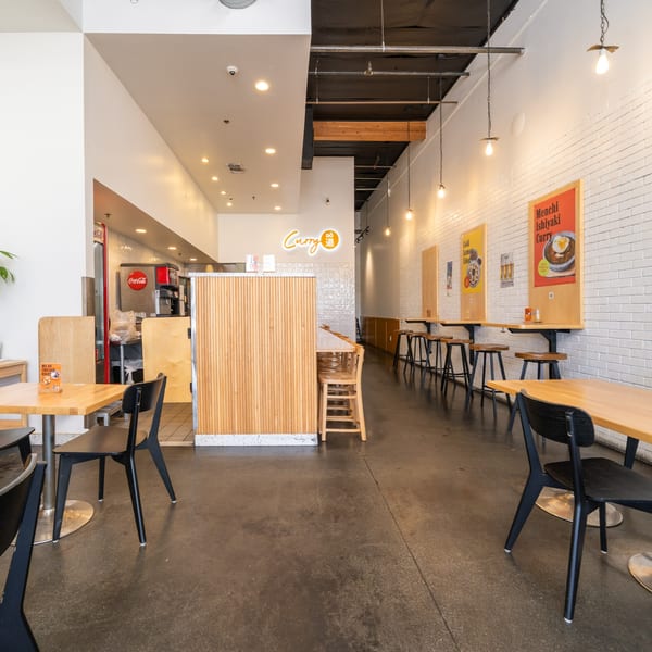 Interior of Curry Do, a Japanese restaurant, with light wood tables, black chairs, and a white brick wall.