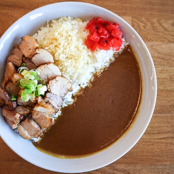 Japanese curry with sliced pork, white rice, tempura flakes, and red pickled ginger in a white bowl.
