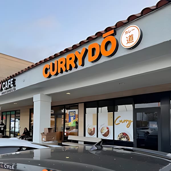 Exterior view of Curry Do restaurant with orange signage and food images on window displays.