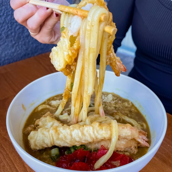 Chopsticks lifting thick udon noodles and tempura shrimp from a bowl of Japanese curry.